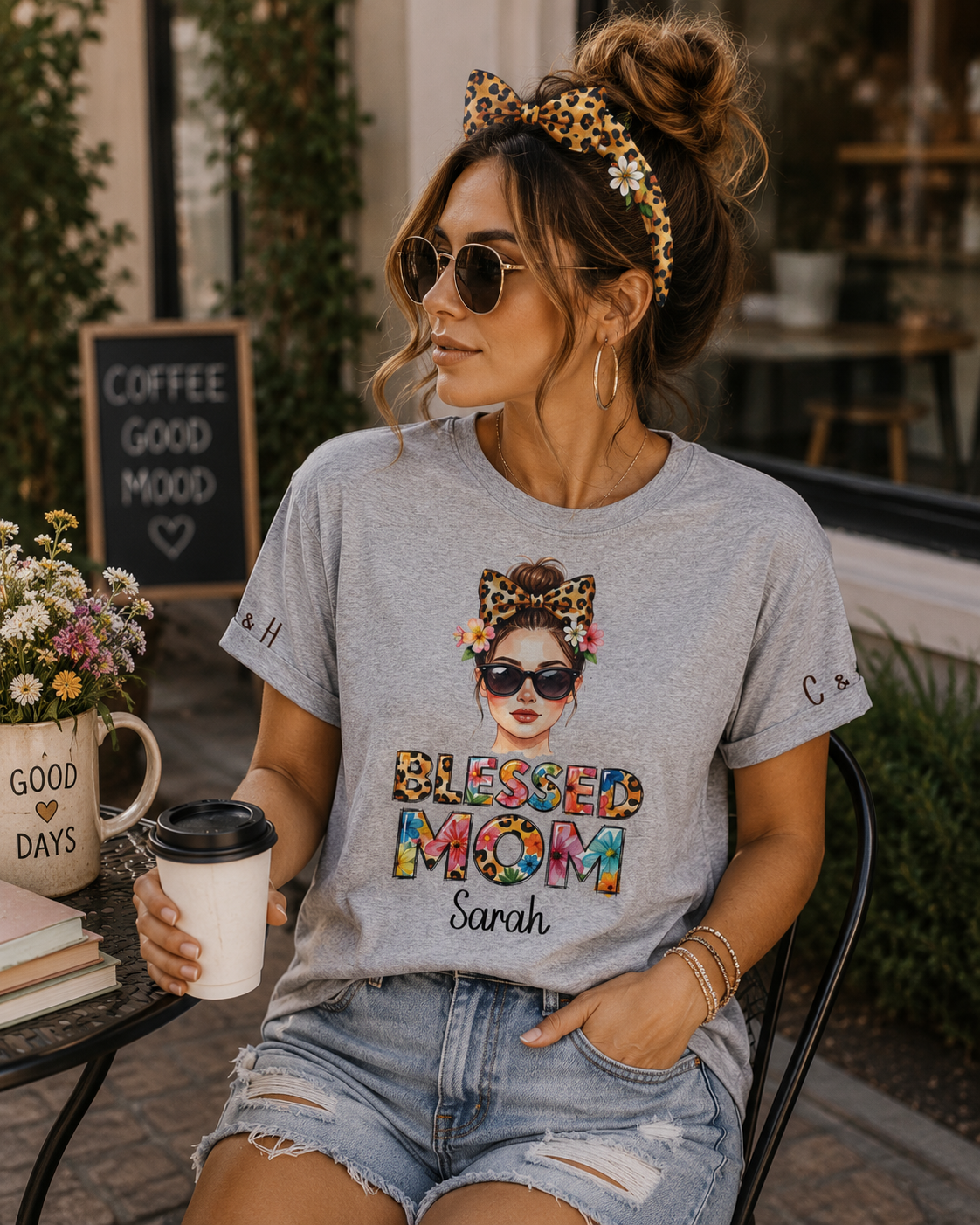 A woman wearing a light gray graphic t-shirt with a colorful 'Blessed Mom' design, styled with denim shorts and a leopard-print hair bow headband, sitting at an outdoor cafe table holding a coffee cup.