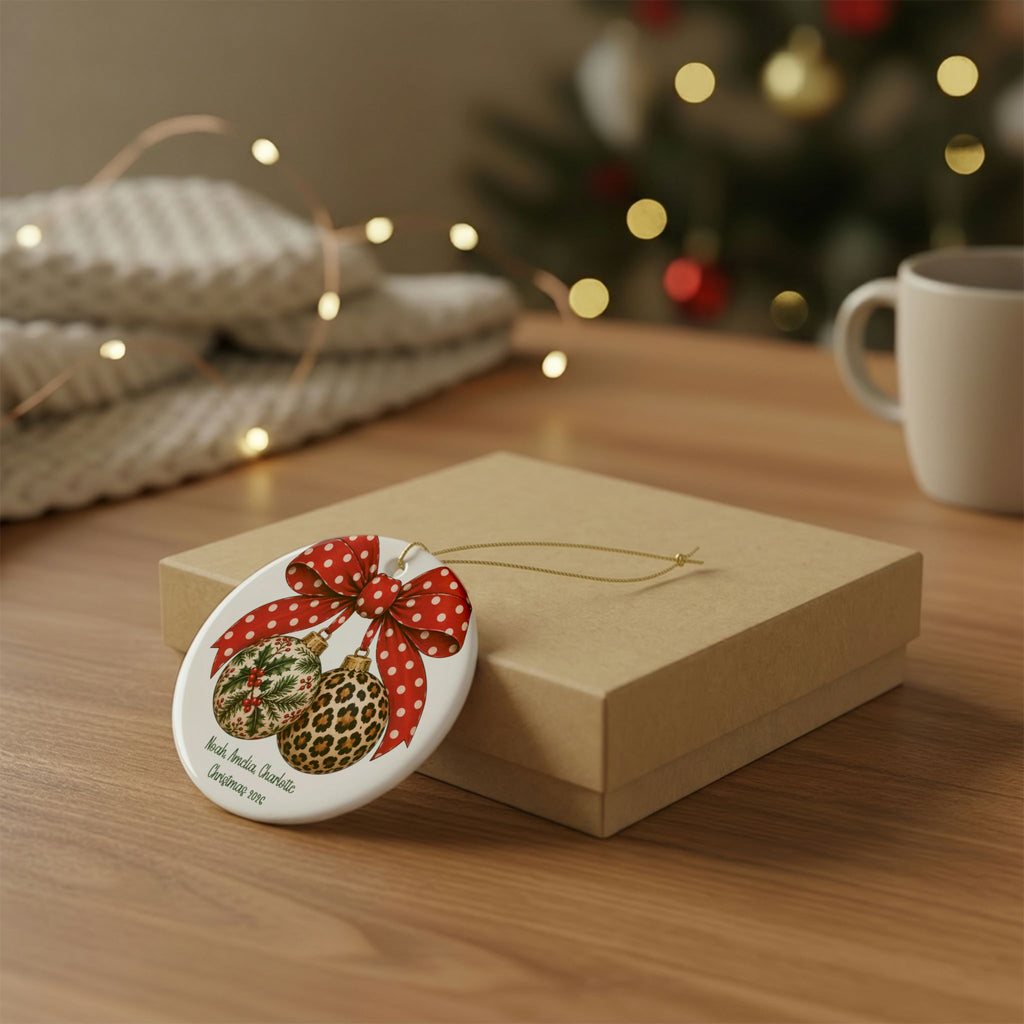 Personalized bow and baubles family ornament resting on a kraft gift box beside a mug and knit throw