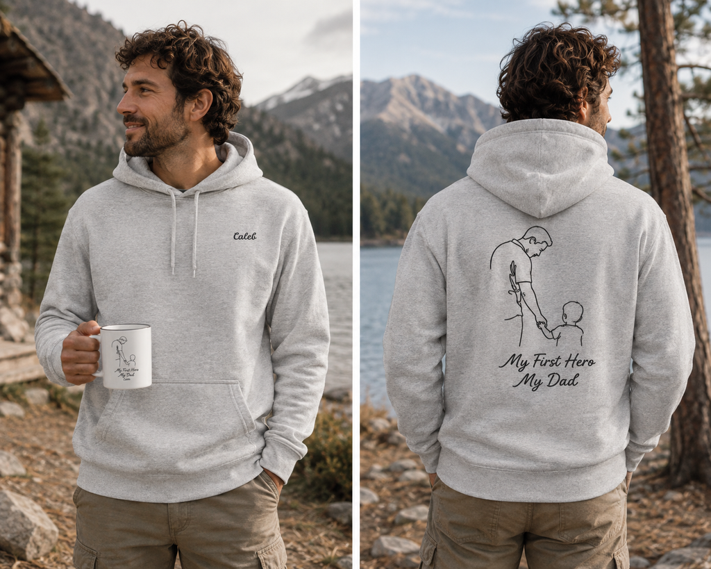 A man wearing a light gray customizable (name) hoodie is shown in a split front and back view against a scenic mountain lake background. The front has a small embroidered 'Caleb' on the chest, and the back features a black line art graphic of a man holding a child's hand with the text 'My First Hero My Dad'.