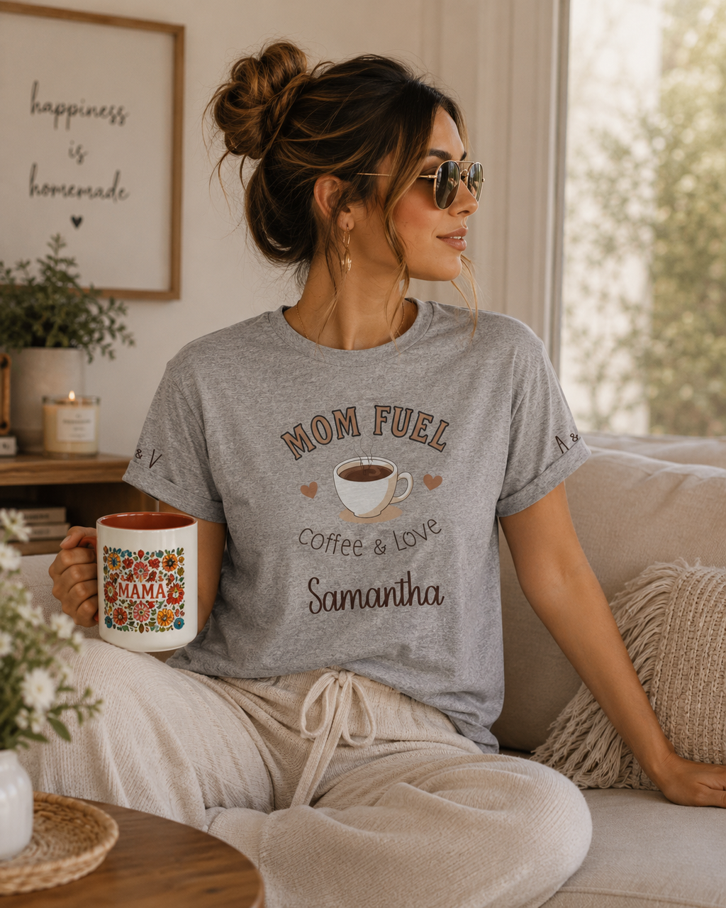 A woman wearing a heather gray graphic t-shirt that reads 'MOM FUEL coffee & love Samantha' sits on a beige couch holding a ceramic mug with a colorful floral pattern and 'MAMA' written on it.