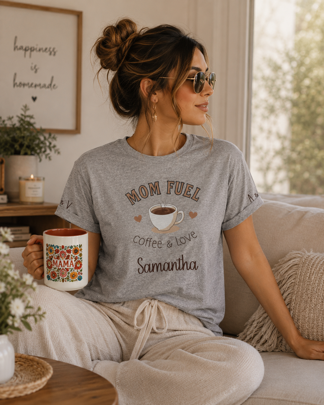 A woman wearing a heather gray graphic t-shirt that reads 'MOM FUEL coffee & love Samantha' sits on a beige couch holding a ceramic mug with a colorful floral pattern and 'MAMA' written on it.