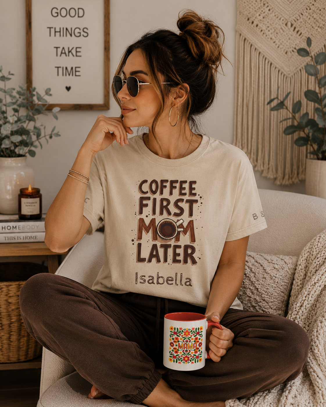 Woman wearing a personalized “Coffee First, Mom Later” T-shirt with the name Isabella, holding a floral mom mug in a cozy boho home setting.
