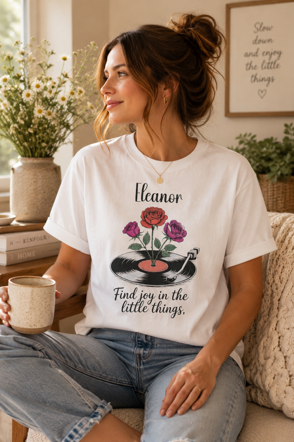 A woman sitting on a couch wearing a white graphic t-shirt with a vintage vinyl record and roses design, holding a mug in a cozy living room setting.