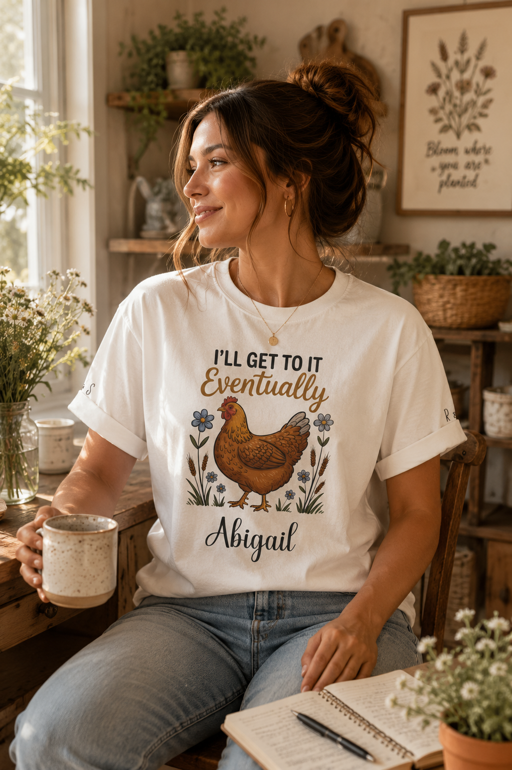 A woman sitting in a cozy room wearing a white graphic t-shirt with a brown chicken illustration and the text 'I'LL GET TO IT Eventually Abigail'. She is holding a mug and wearing blue jeans.