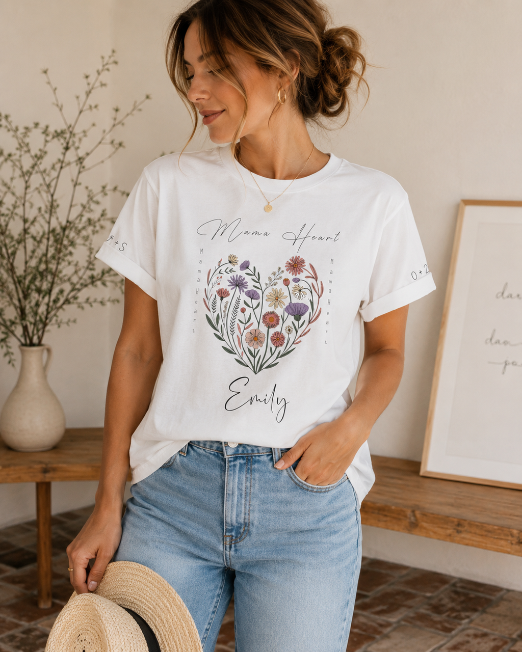 A woman wearing a white graphic t-shirt with a colorful floral heart design and the text 'Mama Heart Emily' printed on the front, paired with light blue jeans in a warm, neutral-toned indoor setting.