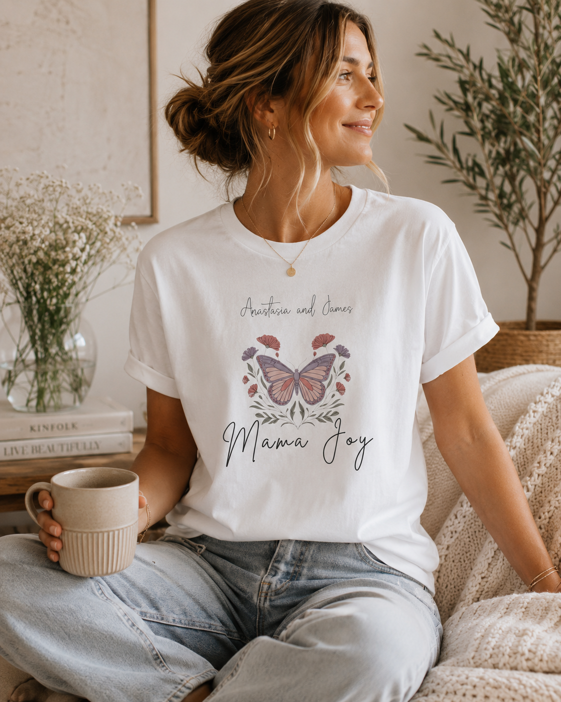 A woman sitting on a couch wearing a white graphic t-shirt with a butterfly design and the words 'Anxiety and Stress' and 'Mama Joy'. She is holding a beige mug in a cozy, well-lit living room setting.