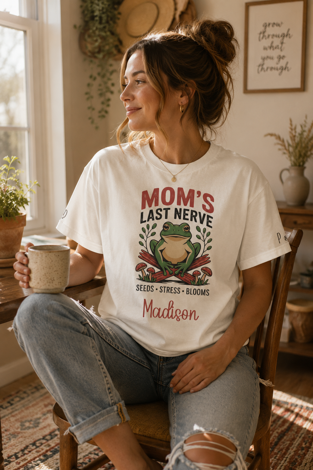 A woman sitting in a rustic room wearing a white graphic t-shirt that reads 'MOM'S LAST NERVE' with a frog illustration and the name 'Madison'.