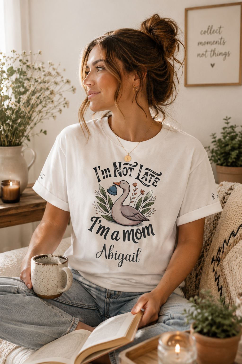 A woman sitting on a couch wearing a white graphic t-shirt with a swan illustration and the text 'I'm Not Late I'm a mom Abigail'. She is holding a mug and an open book in a cozy, sunlit living room setting.