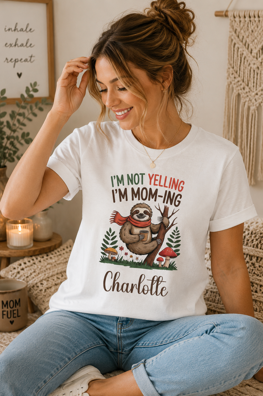 A woman sitting indoors wearing a white graphic T-shirt with a sloth illustration and the text 'I'M NOT YELLING I'M MOM-ING' and 'Charlotte,' paired with light blue jeans.