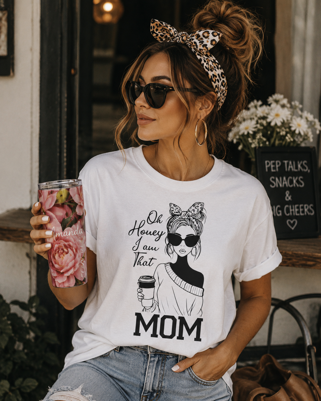 A woman wearing a white graphic t-shirt with a black print of a woman in sunglasses and the text 'Oh Honey I Saw That MOM', paired with light-wash jeans and a leopard-print headband, holding a floral tumbler in a cafe setting.