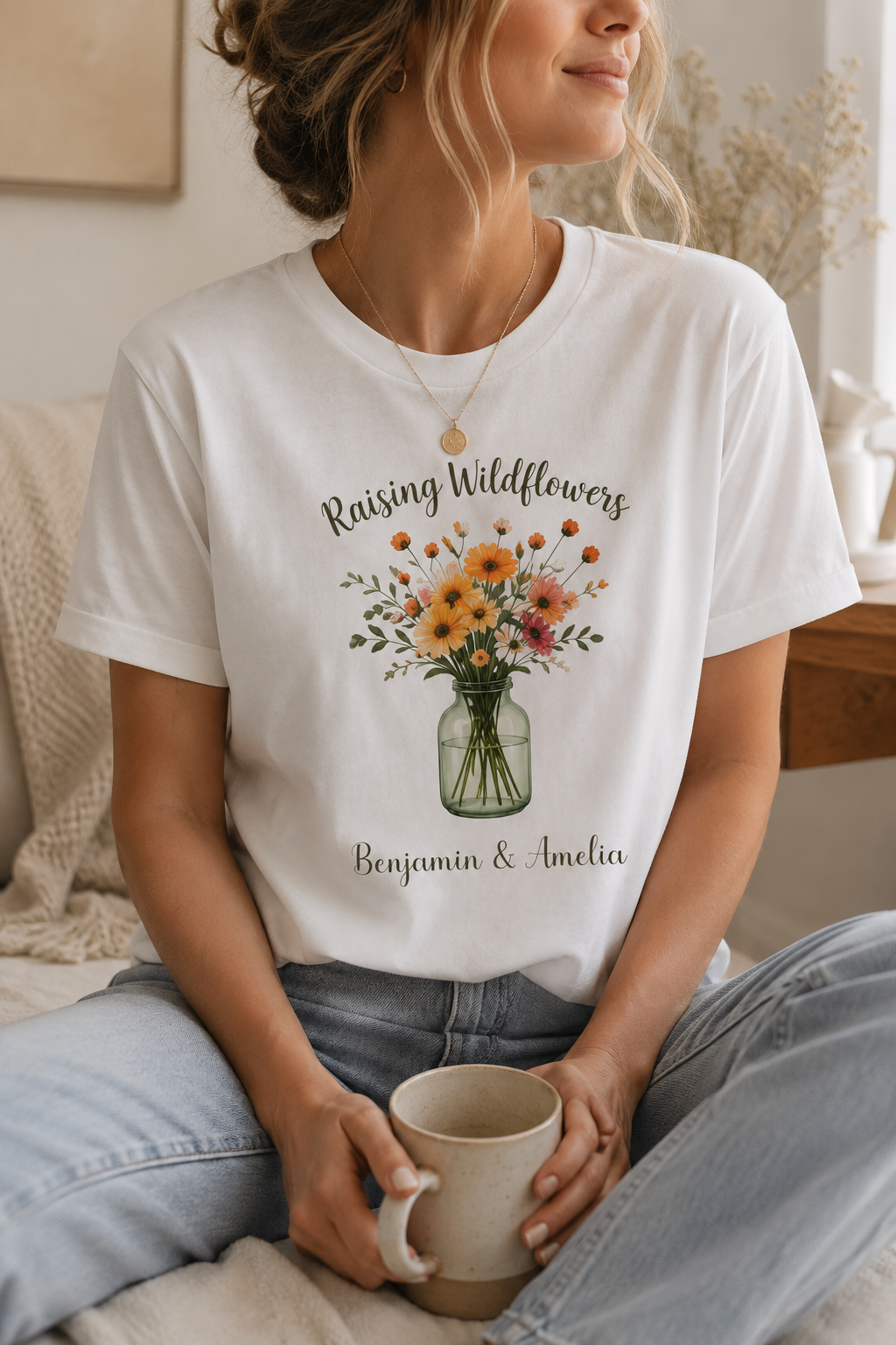 A woman wearing a white graphic t-shirt with a 'Raising Wildflowers' floral print and the names 'Benjamin & Amelia' in script, sitting in a cozy indoor setting and holding a mug.