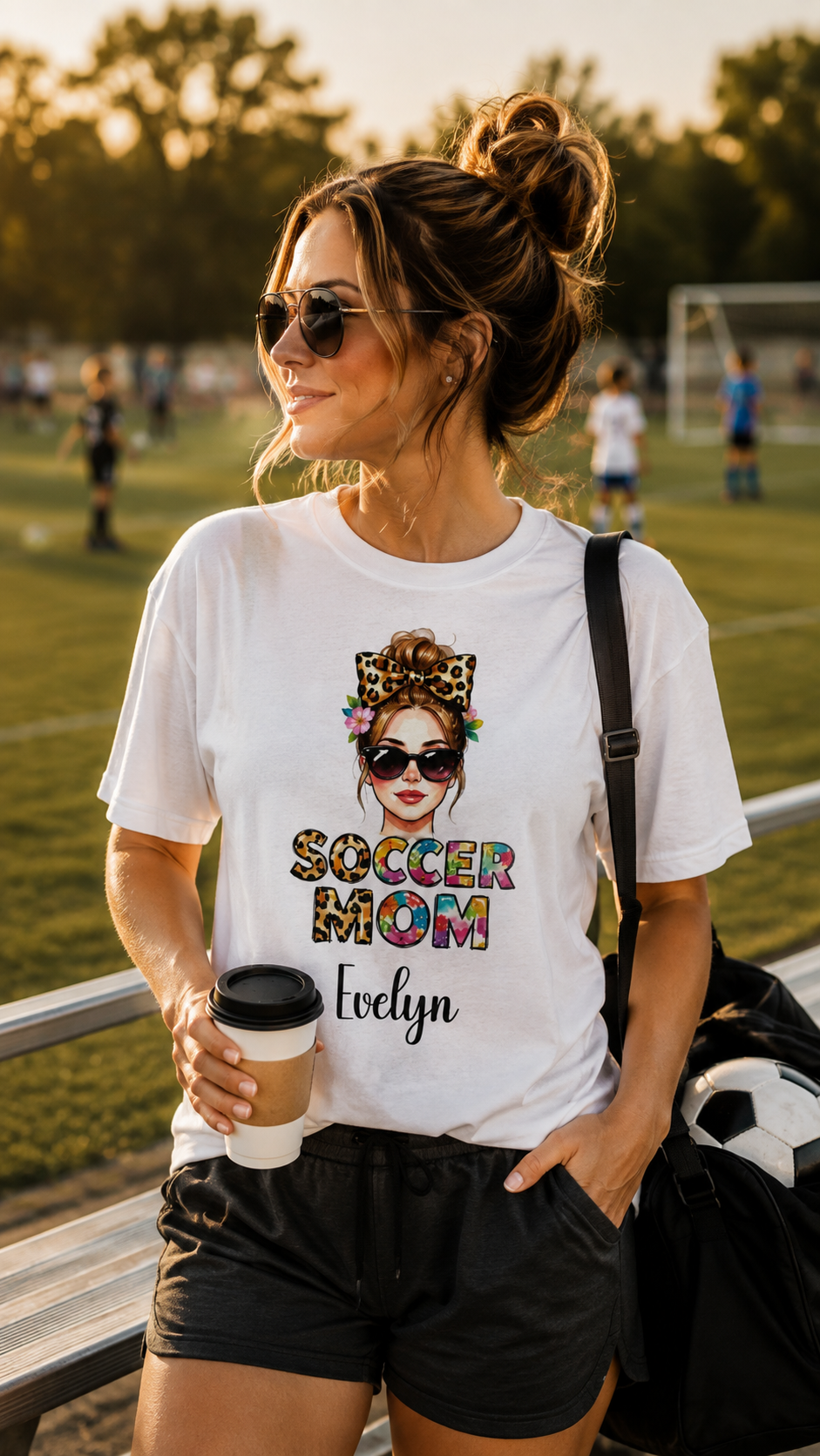 A woman wearing a white graphic t-shirt with a colorful 'Soccer Mom' design and the name 'Evelyn' printed on the front, standing on a soccer field at sunset.