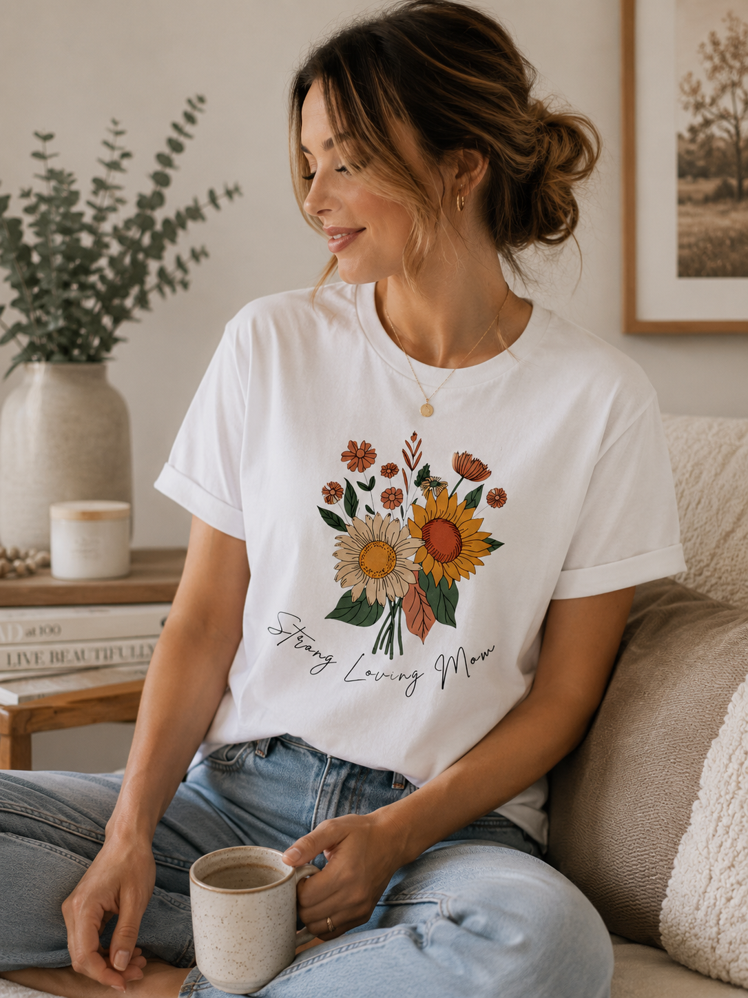 Woman wearing a personalized “Strong Loving Mom” white T-shirt with a colorful floral design, relaxing at home with a coffee mug.