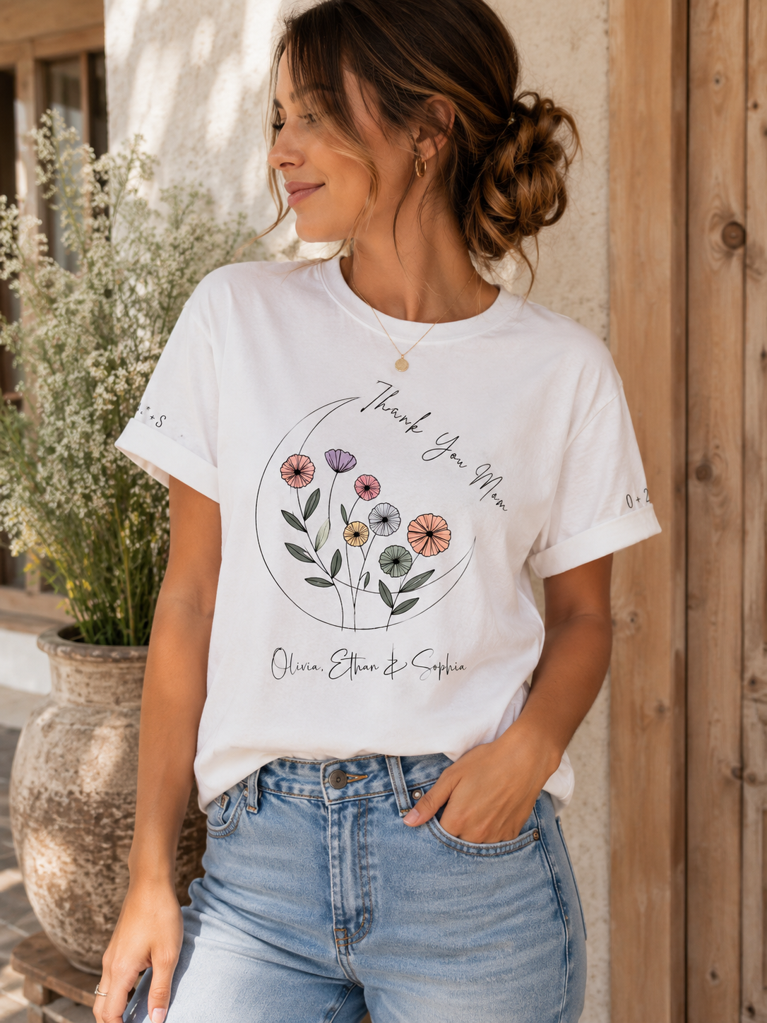 A woman wearing a white graphic t-shirt with a colorful floral design and the text 'Thank You Mom' and 'Olive Elman & Sophie', paired with light blue jeans, standing in front of a rustic wooden door.
