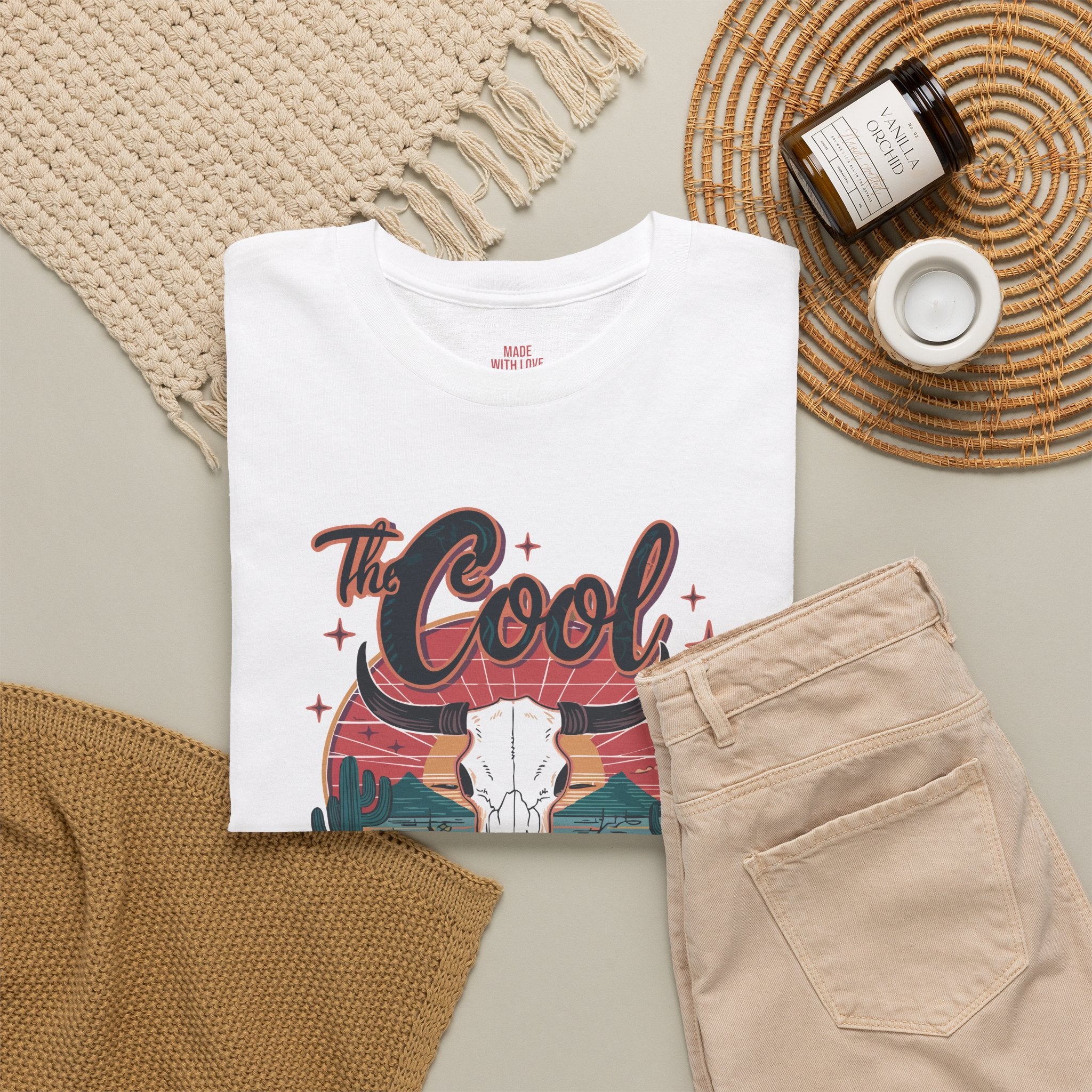 A white graphic T-shirt laid flat on a neutral surface, styled with beige pants, a woven blanket, a candle jar, and a small cup. The shirt features a western-themed graphic with the words “The Cool” above a bull skull and desert landscape.