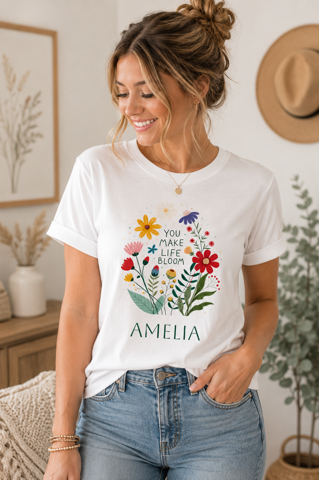 A woman wearing a white graphic t-shirt with a colorful floral design and the text 'YOU MAKE LIFE BLOOM' above the name 'AMELIA', paired with blue jeans in a cozy indoor setting.