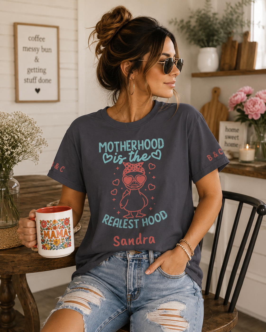A woman wearing a dark gray graphic t-shirt with a 'Motherhood is the Realest Hood' design, sitting in a cozy kitchen setting and holding a mug.